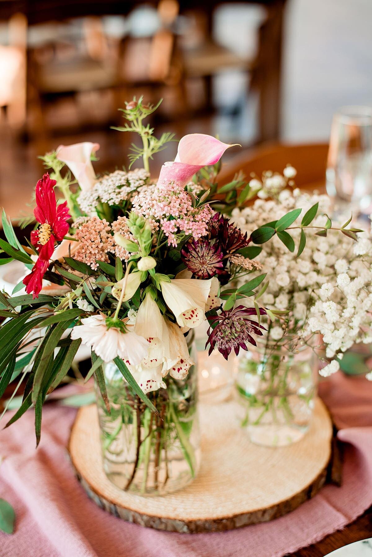 Sitting on a wood slice, clear glass bud vases are clustered together with a variety of Tennessee wild flowers in red, white, pink, ivory and burgundy.