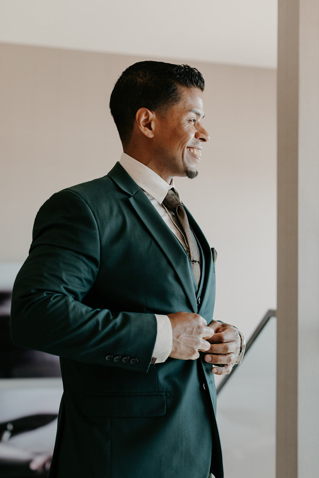 The groom, dressed in a tailored green suit and dark tie, smiles confidently as he fastens his suit jacket, ready for the wedding ceremony.
