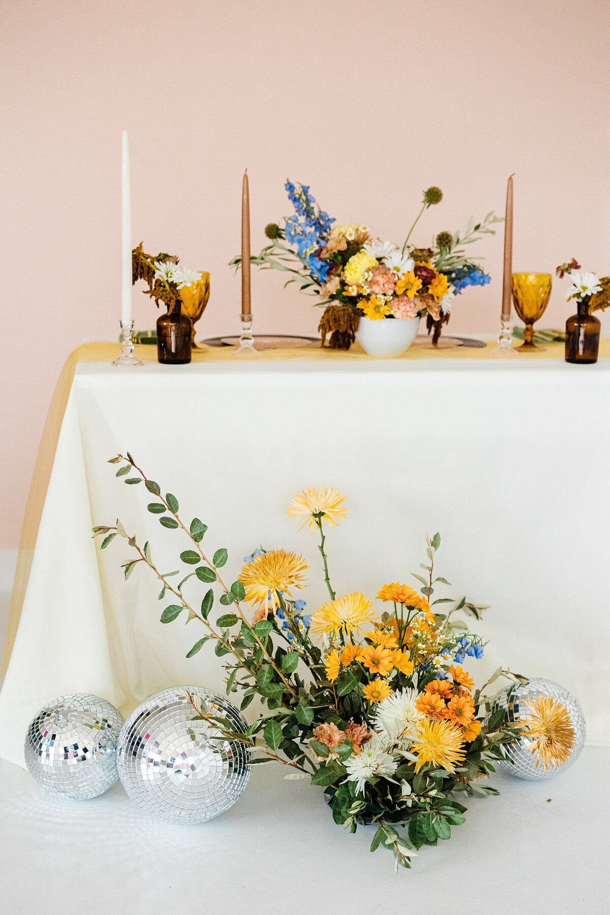 Detail shot of a reception table at a wedding in Dallas, Texas. The table has a long, white table cloth, place settings, many candles, and a bouquet of colorful flowers as the centerpiece. In front of the table is a similar but larger floral arrangement and multiple disco balls of varying sizes.