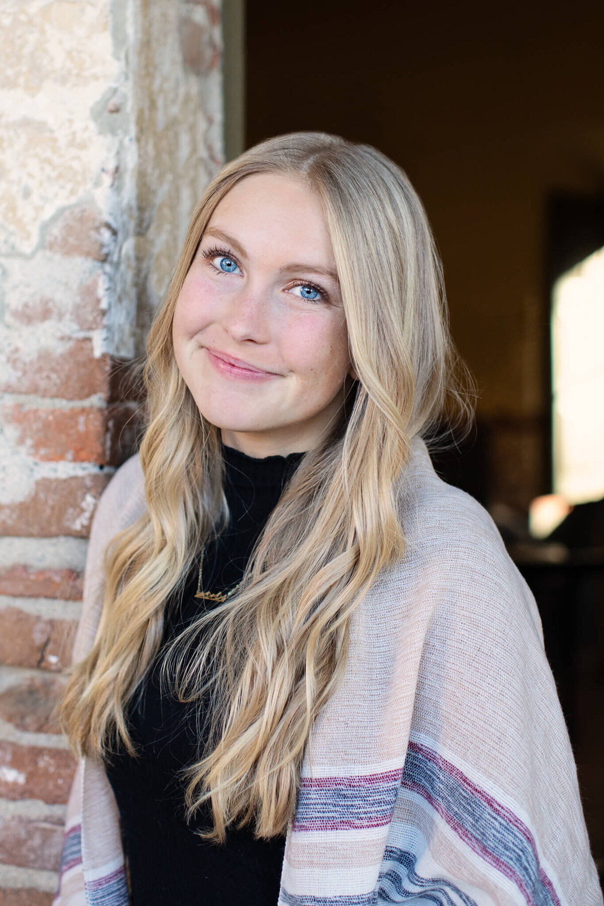 A close-up of a beautiful woman with bright blue eyes and long, platinum blonde hair, wearing a black turtleneck and a grey and beige cardigan, leaning against a brick column, smiling at the camera with just her lips.