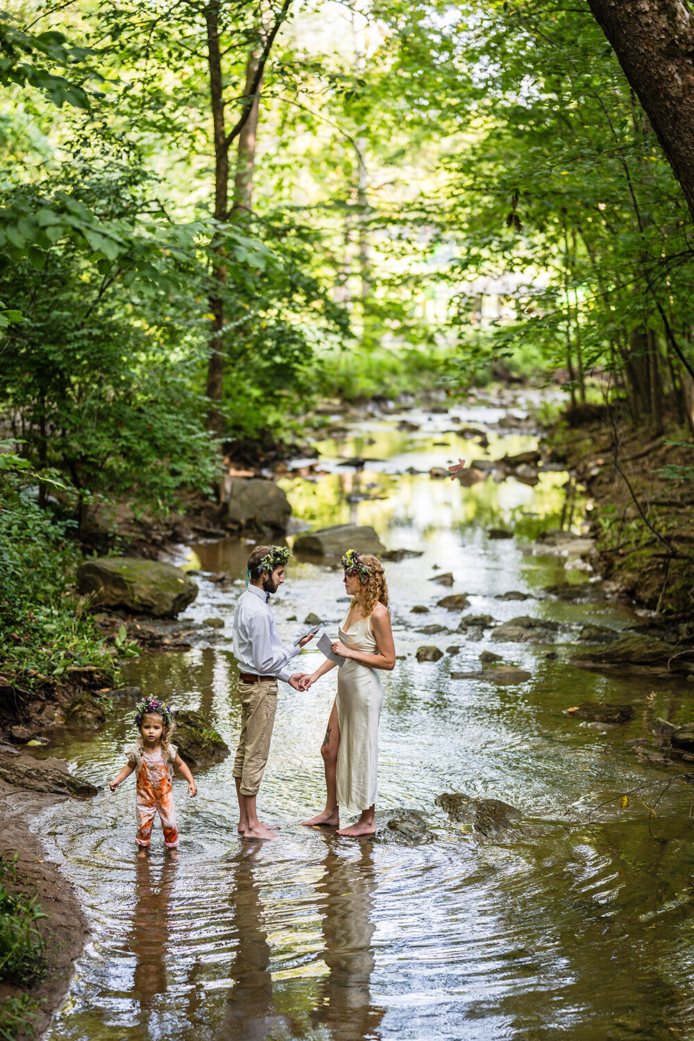 A couple on their elopement day face one another and read their vows while standing in the creek at Fishburn Park. Their child can be seen behind the groom looking out in one direction as she steps in the water.