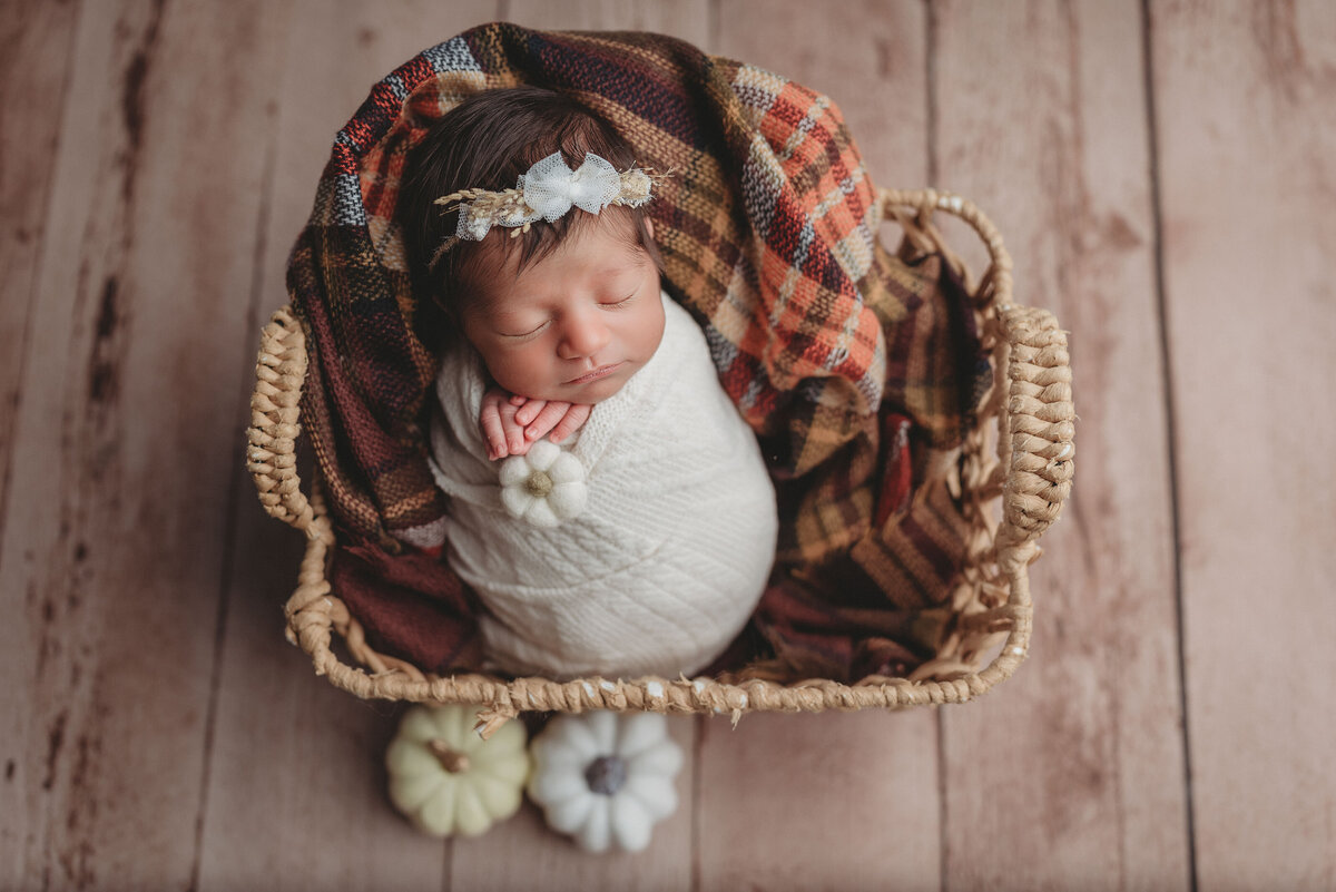 Baby girl in white swaddle asleep in basket with fall pumpkin decorations and plaid scarf