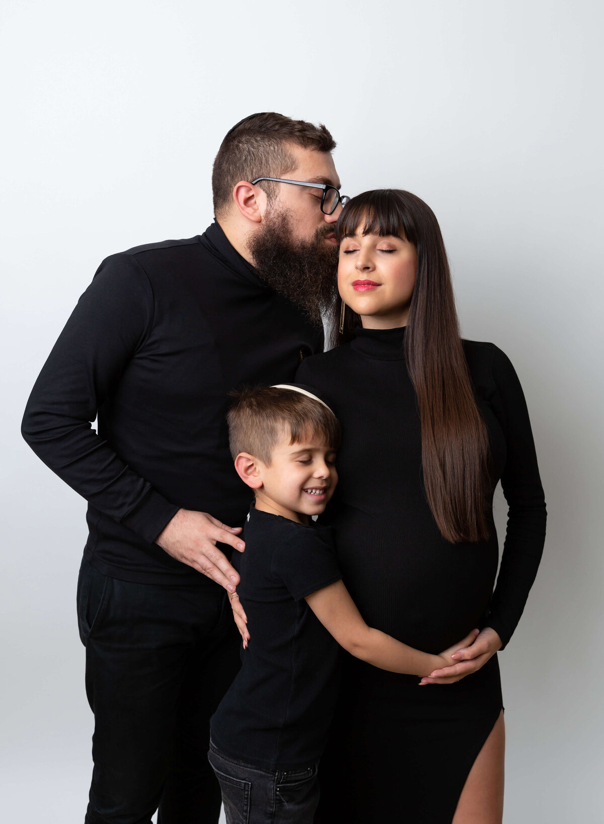 A mom-to-be stands in a studio in a black maternity gown with a hand on her head and belly after a pregnancy photoshoot in NYC