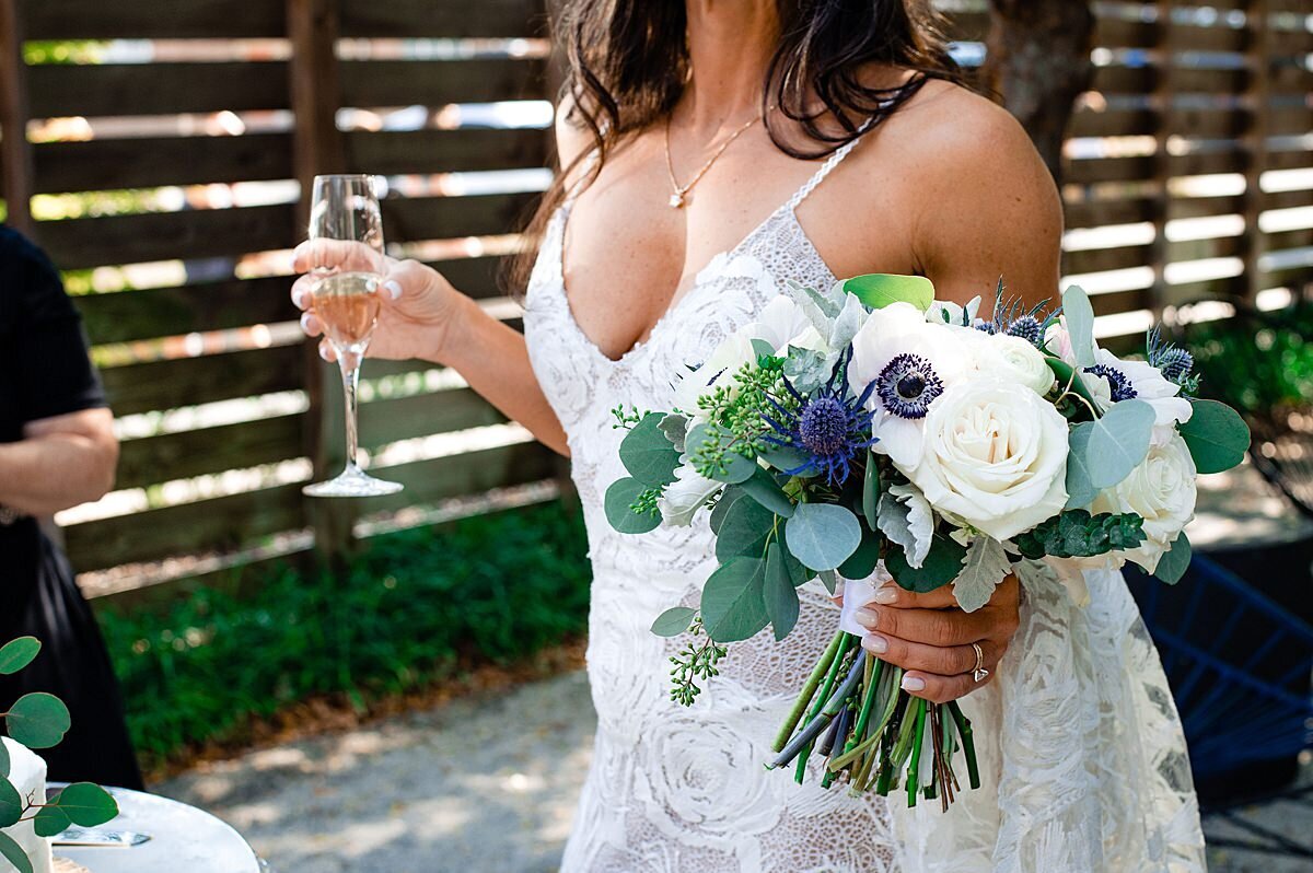 The bride, wearing a v-necked lace boho wedding dress holds a champagne flute and her bouquet of anemone, blue thistle, white and ivory roses with seeded and silver dollar eucalyptus at her Nashville elopement.