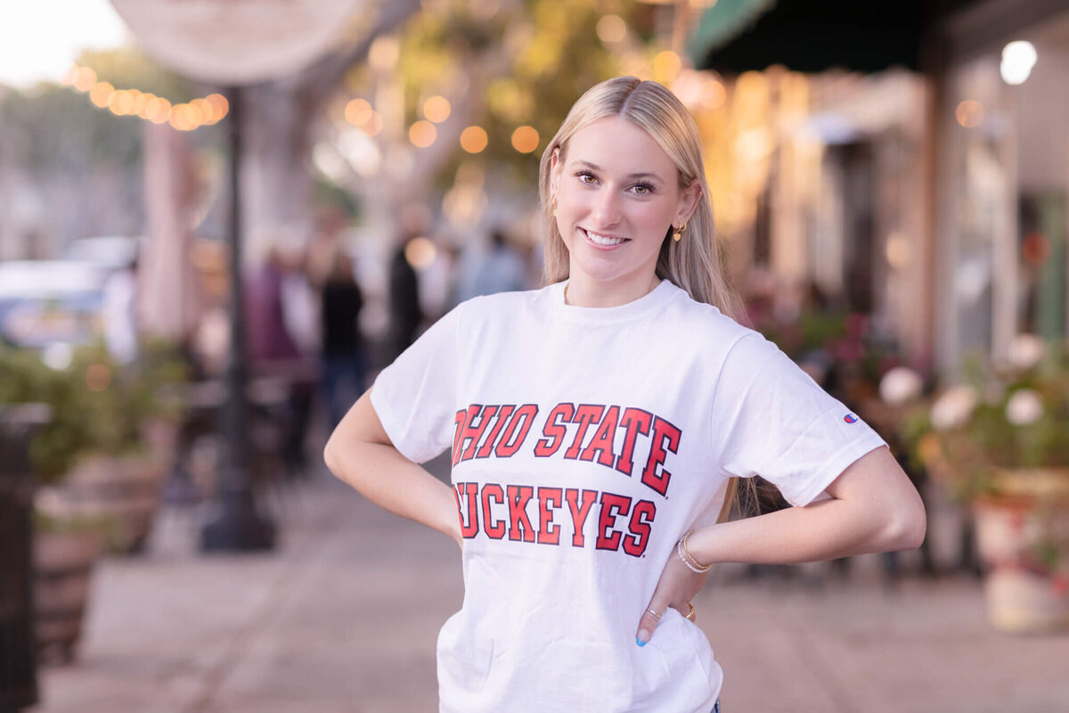 A person wearing a white Ohio State Buckeyes t-shirt stands smiling with their hands on their hips. They are on a sidewalk with blurred lights and shops in the background.