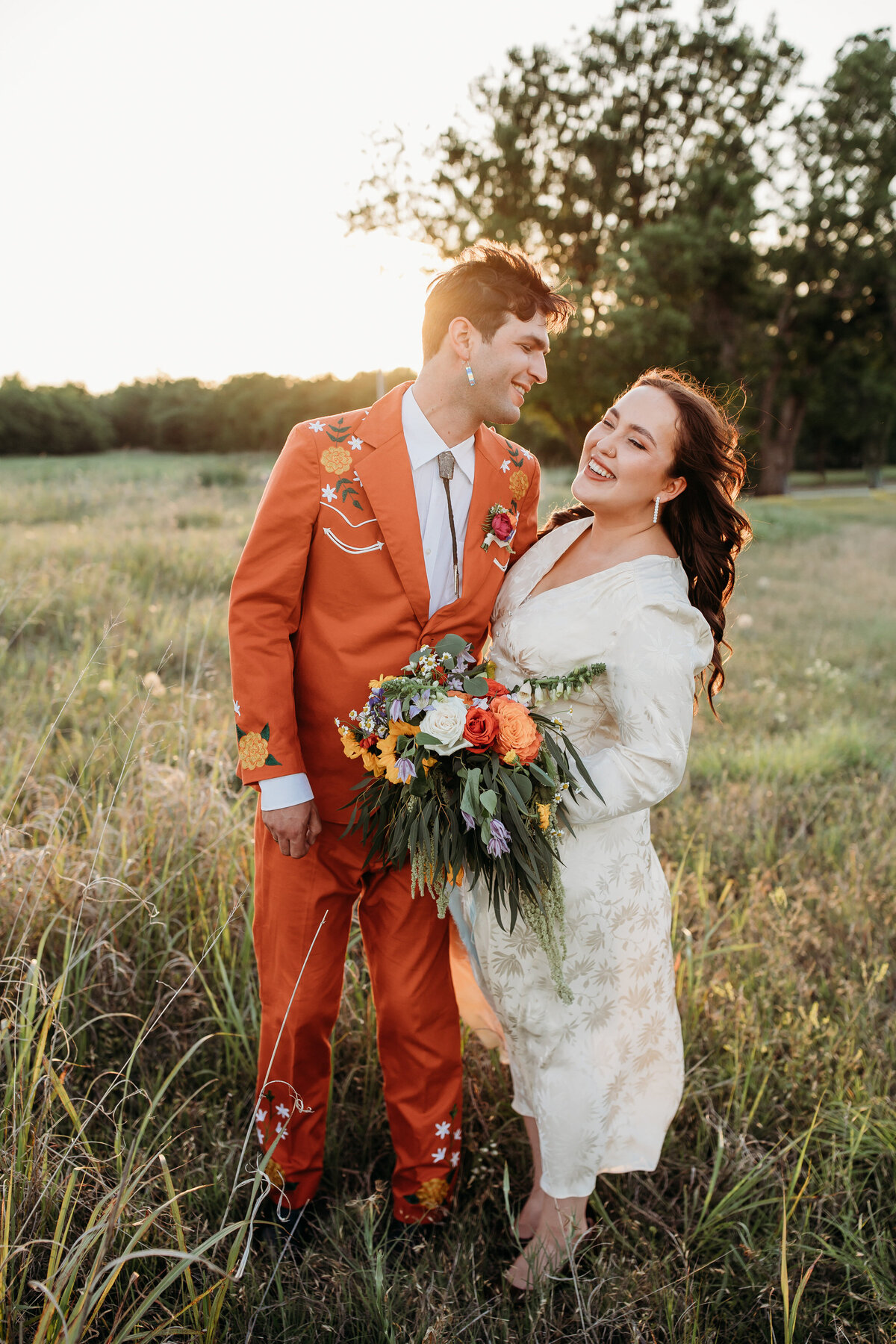 Couple in a sunset field, with the partner in a vibrant orange suit and the other in a white dress, both smiling lovingly