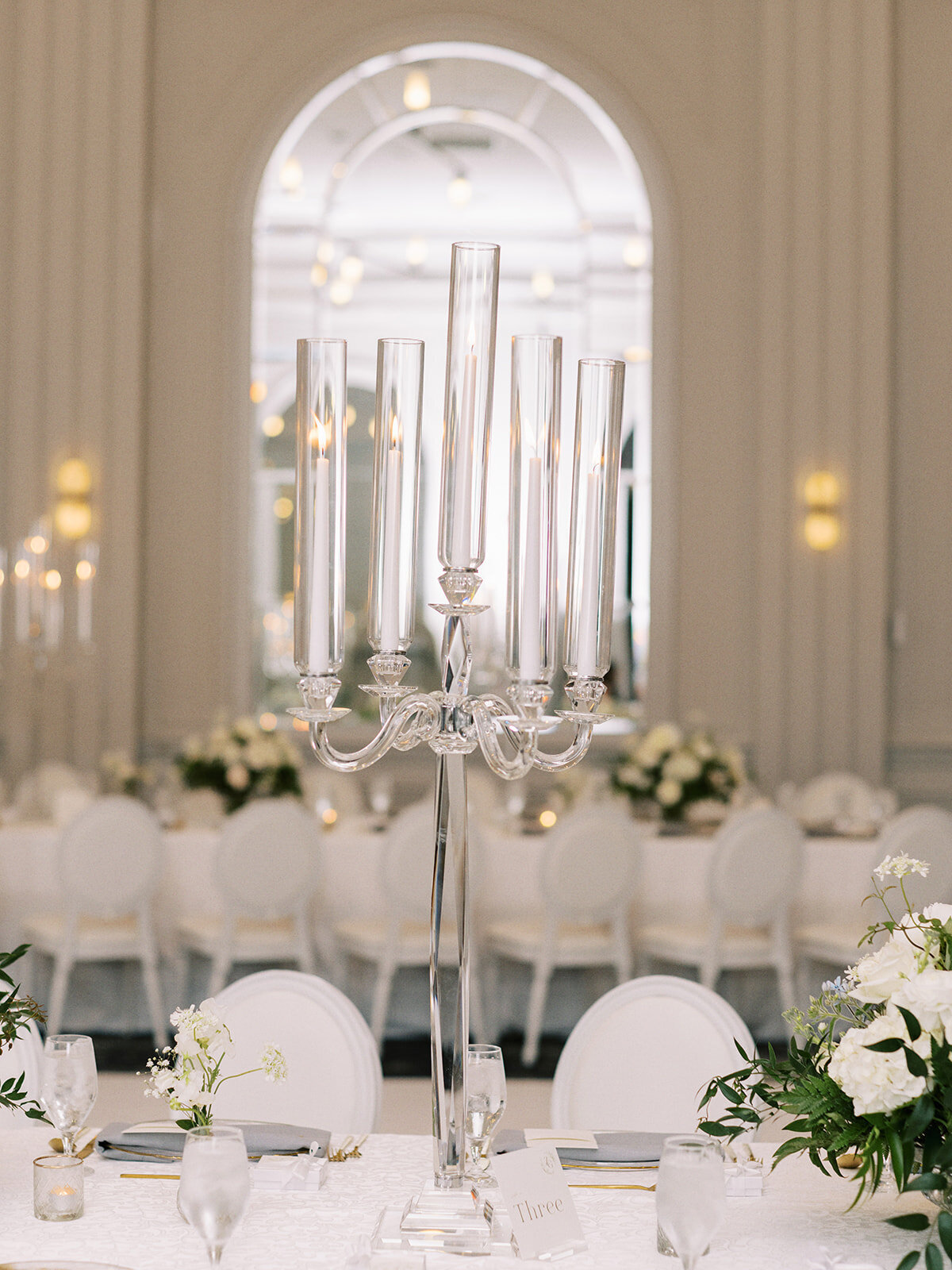 A tall, elegant candelabra with lit candles serves as a centerpiece on a formal dining table, surrounded by white chairs and greenery in a large, well-lit room featuring an arched mirror in the background—a perfect setting for a classic Calgary wedding.