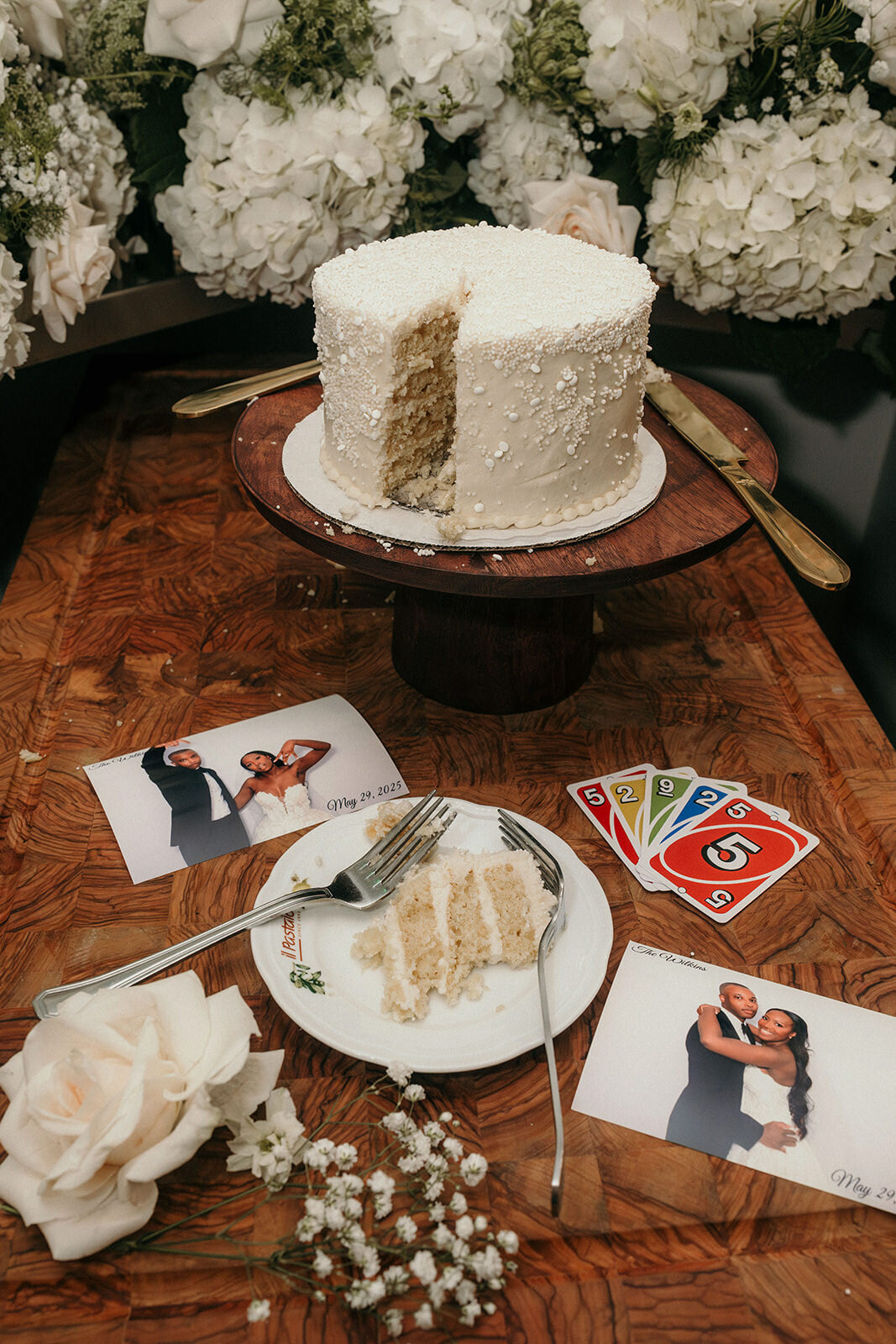 First slice of the wedding cake on a table, surrounded by pictures of the newly weds and some UNO-cards.