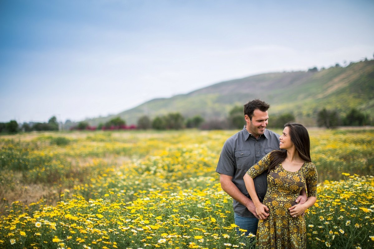 Bride to be looks back as her fiance holds her from behind in an open field full of yellow flowers