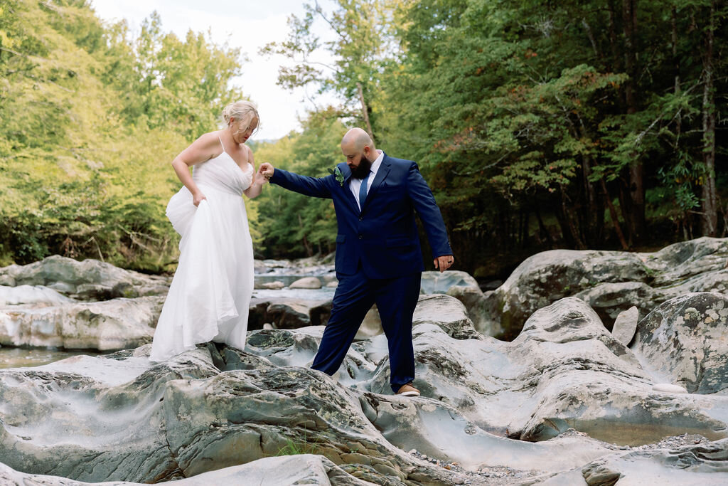 Groom helping the bride carefully step across smooth river rocks at Greenbrier during their eloping to Gatlinburg celebration, surrounded by natural forest and flowing water.
