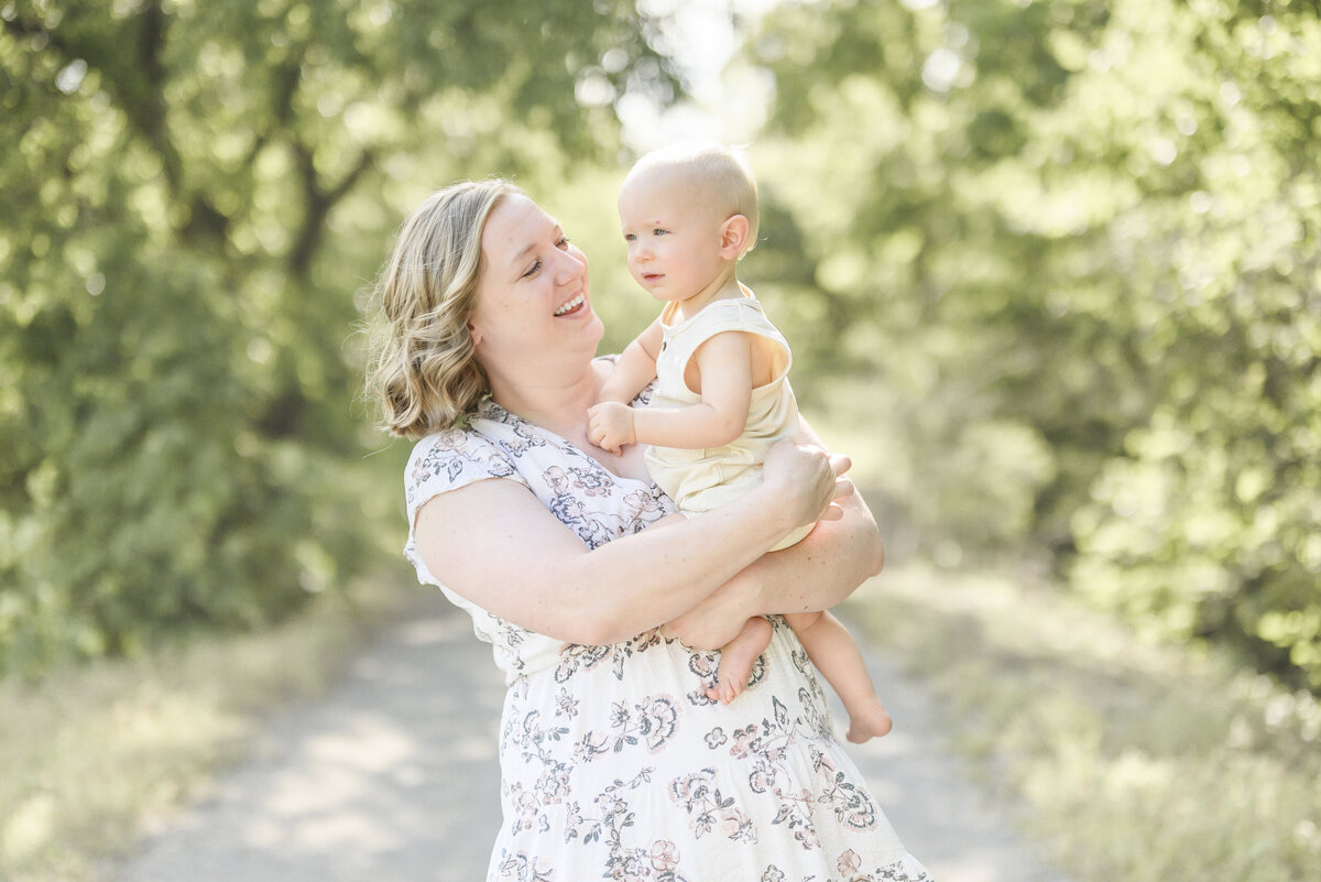 Mom holding her smiling baby in a light floral dress on a sun-dappled wooded path during a warm summer family session.