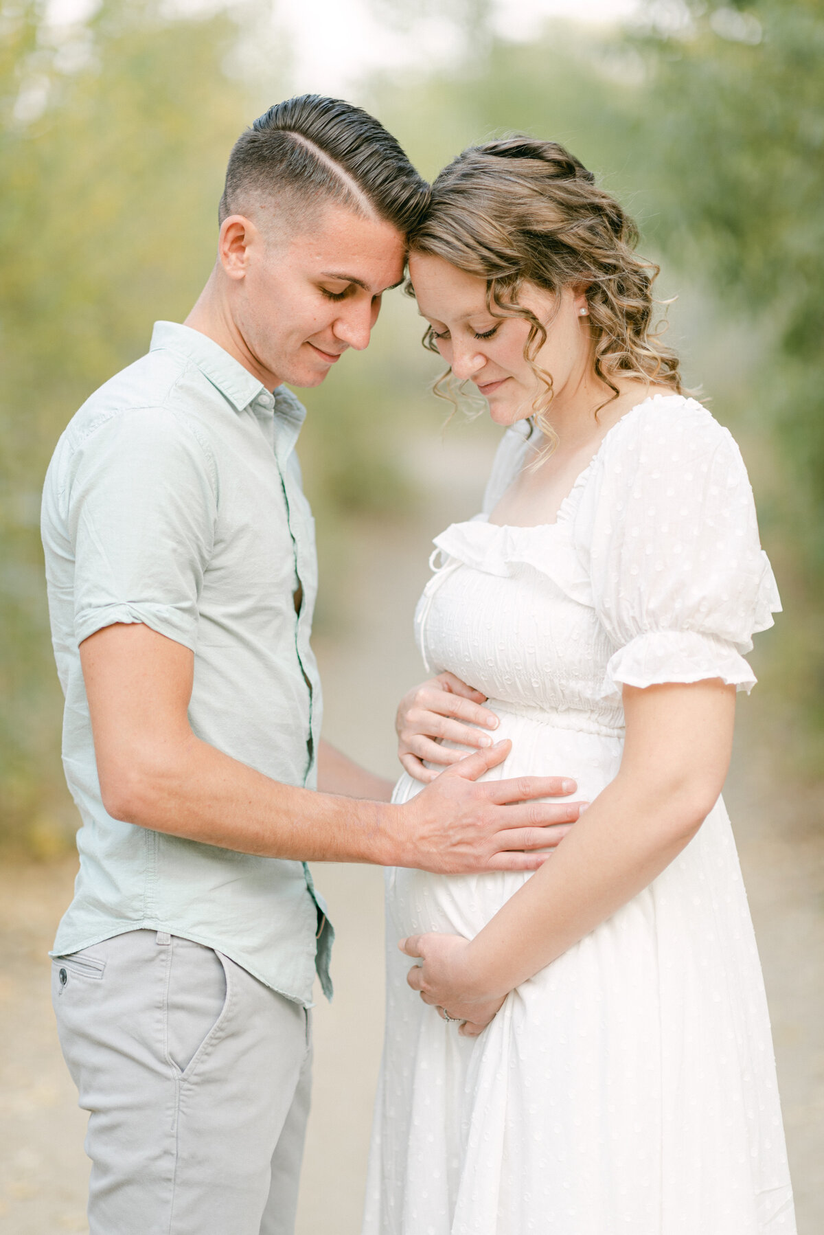 Portrait of a man and a pregnant woman outdoors leaning into each other looking down with their hands placed on her stomach.