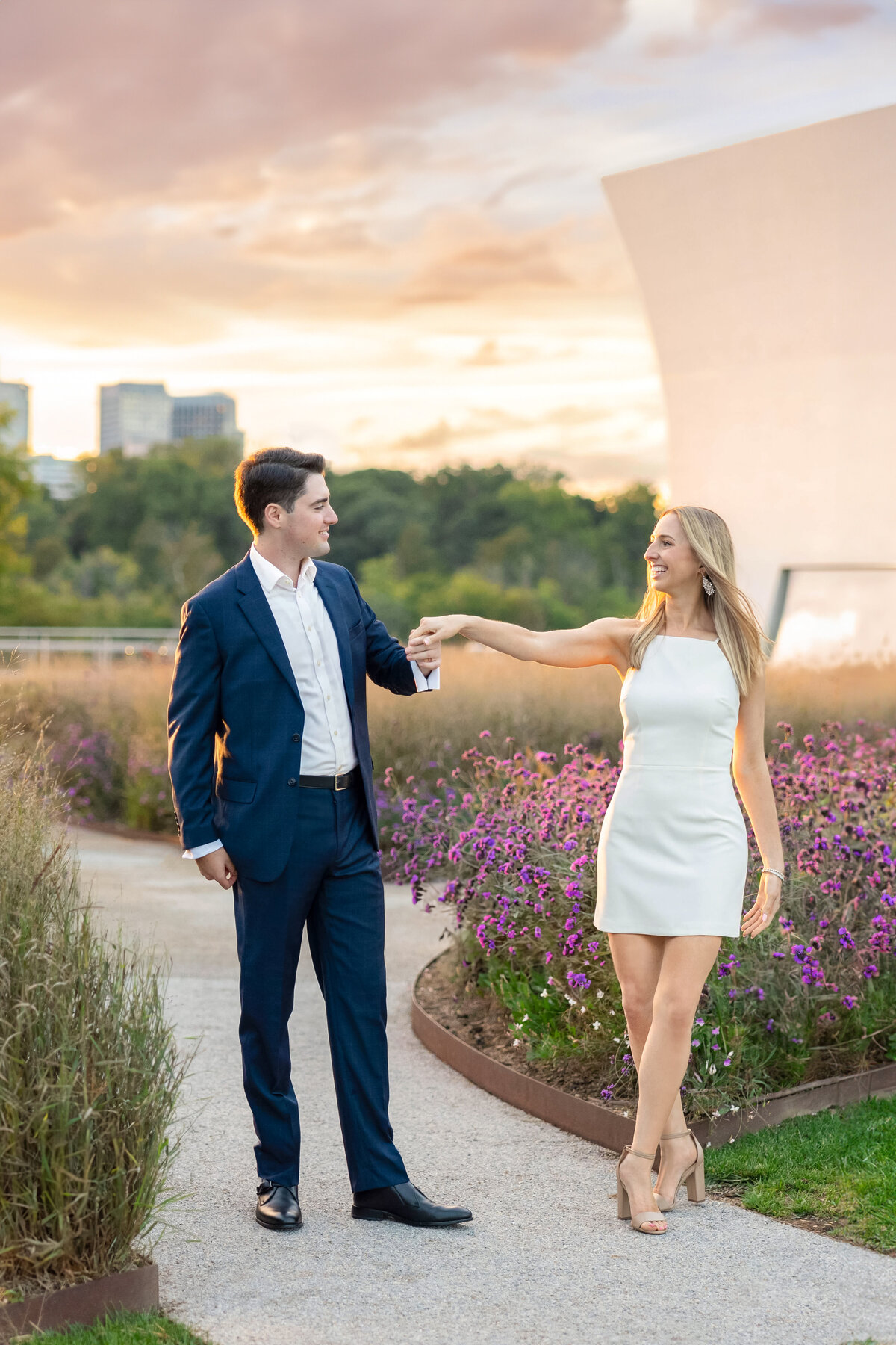 A couple dances on a pathway lined with purple flowers at sunset. The man wears a navy suit, and the woman wears a white dress and heels. In the background, trees and modern buildings are visible under a colorful sky.