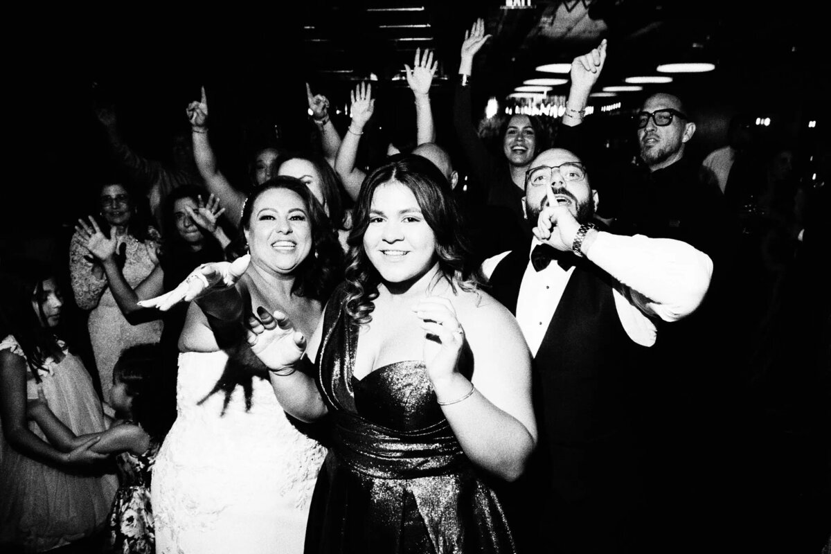 A joyful group of people, including a bride in a wedding dress, pose and celebrate together at a lively indoor event, beautifully captured by an NJ wedding photographer. Many are smiling, cheering, and raising their hands in excitement.