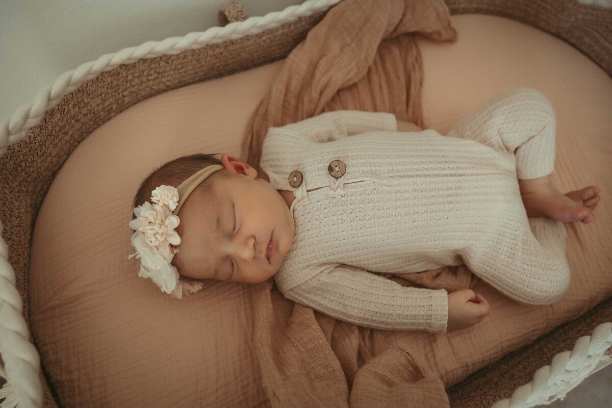 sweet baby girl is sleeping in a basket. she is wearing a flower headband and a white onesie. She is laying on a dusty pink blanket