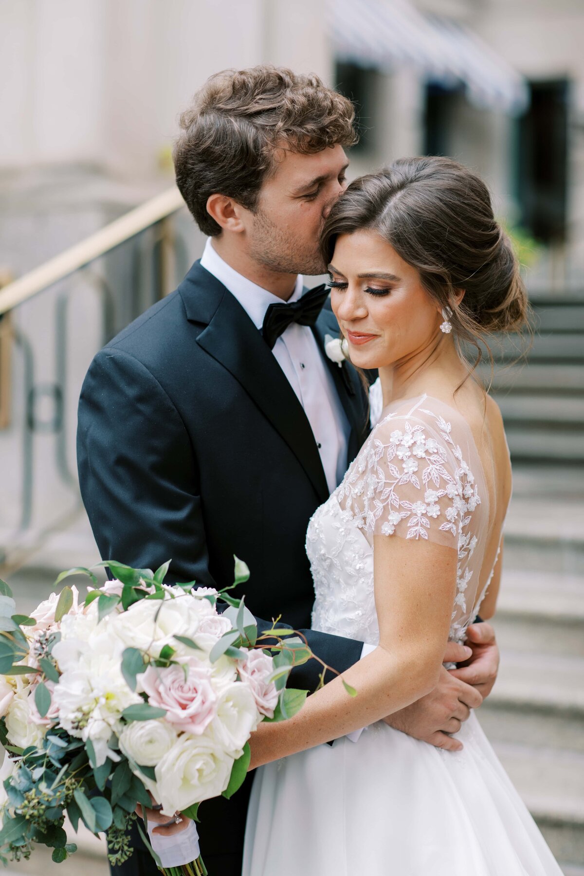 Handsome groom in a black tie tux kissing the temple of a gorgeous brunette bride in a wedding gown holding a white rose bouquet on the steps of The Willard Hotel