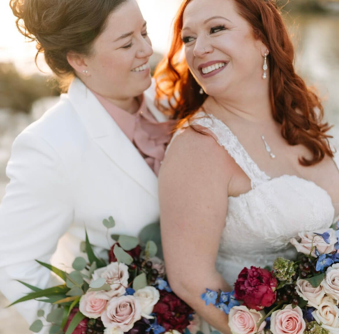 LGBTQ+ wedding photo of two brides smiling—one in a lace gown with red curls and bold lipstick, the other in a white suit with soft glam makeup. Mistique Makeup provides inclusive wedding day hair and makeup services in Austin, Dallas, and San Antonio. Mobile bridal beauty team available for all couples.