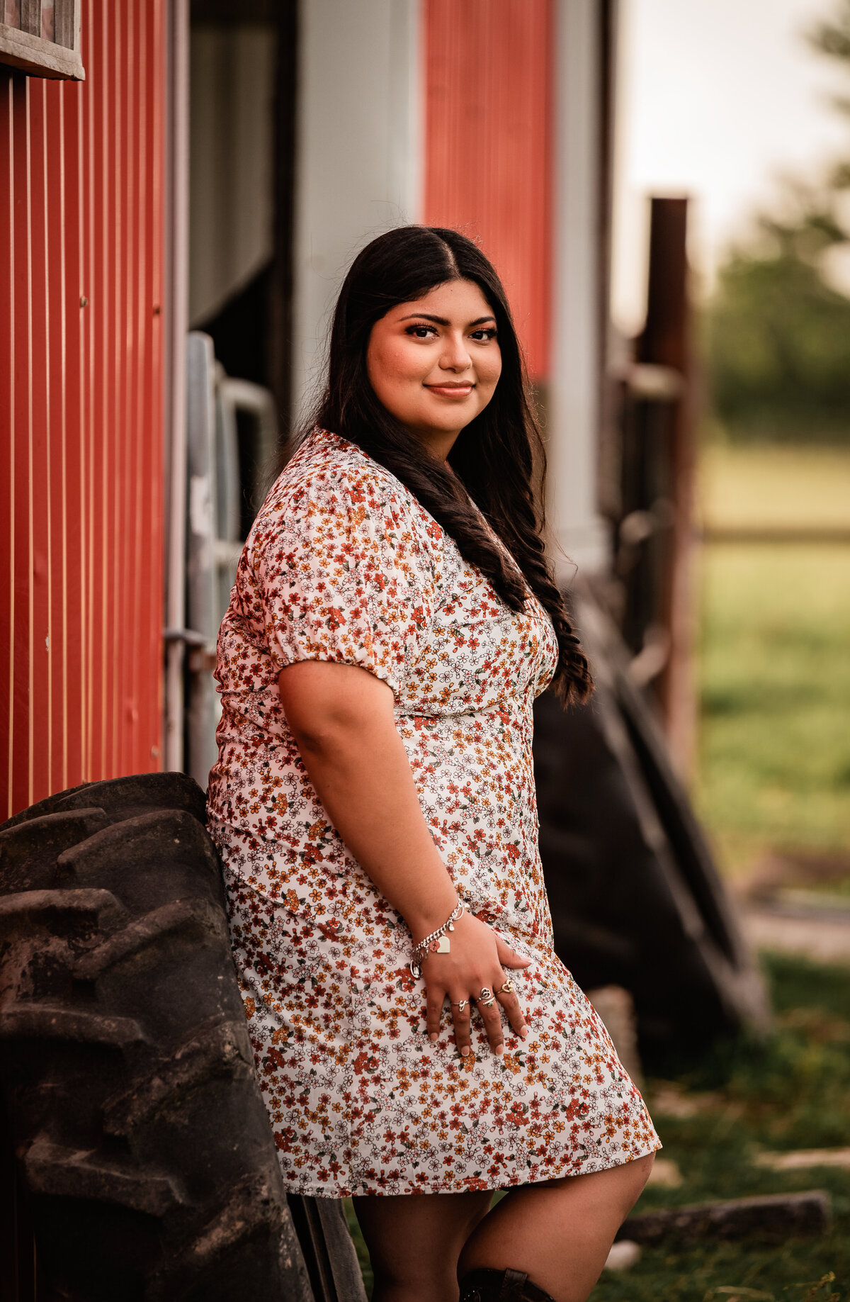 A girl wearing a floral dress leans against a tractor tire and smiles at the camera for her senior portrait.