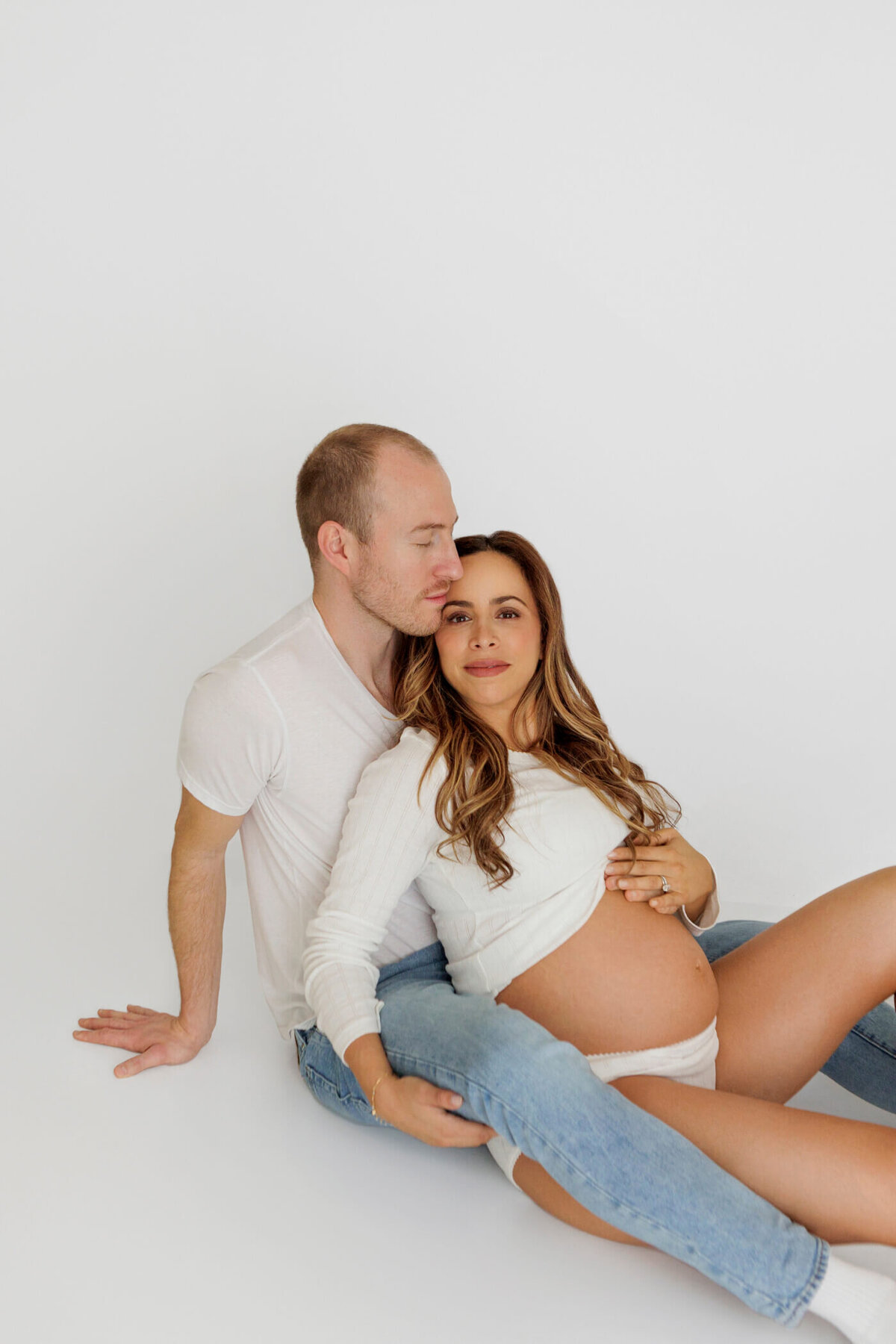 A pregnant woman sits on the floor, leaning back against her partner, who lovingly kisses her on the forehead. She wears a white long-sleeve top and underwear, with her bare baby bump visible. Her partner, dressed in a white t-shirt and jeans, wraps his arm around her, supporting her as they sit together. The couple shares an intimate and peaceful moment, captured in a bright, minimalist setting that emphasizes their connection and anticipation of the new arrival.