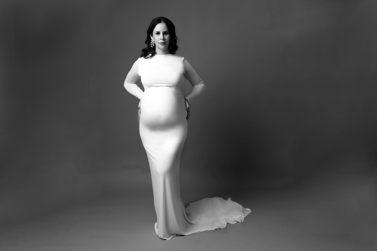 A black and white portrait of a woman standing wearing a white gown with tail and showing her pregnant belly