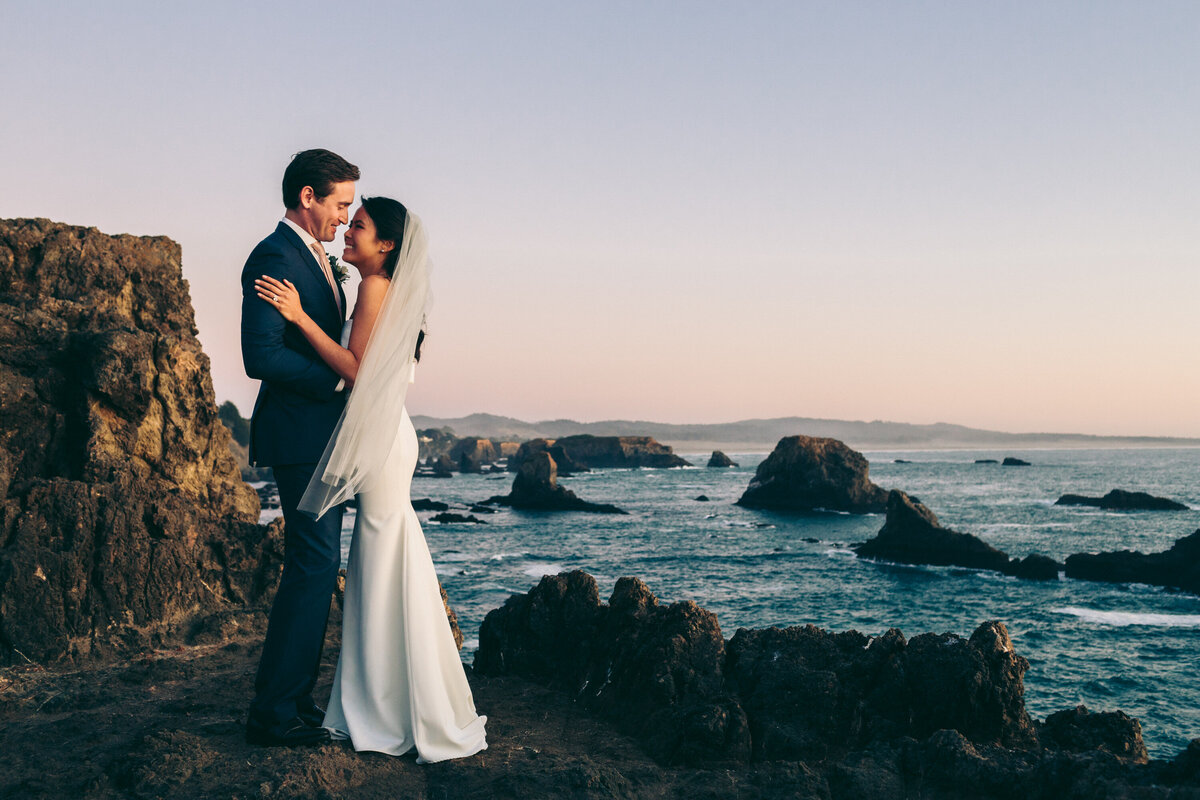 A photograph in color of Melissa and Doug on their wedding day at The Inn at Newport Ranch in Fort Bragg, California. The portrait of the bride and groom is taken at sunset. The landscape is the ocean and rugged coastline or rocky cliffs and tall rocks in the blue water. The sun has just gone down so the sky is pastel in color and a soft glow shines on the couple. They are facing each other in an embrace while smiling and laughing. Wedding photography taken by Stacie McChesney/Vitae Weddings.