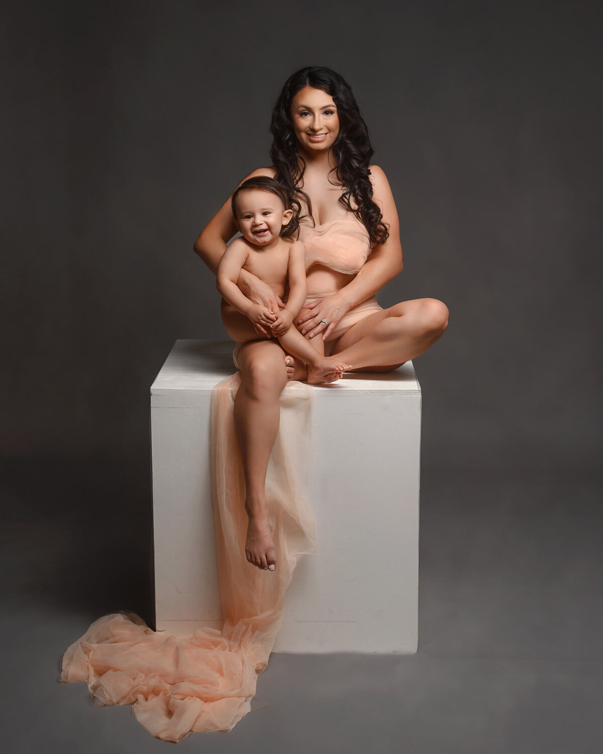 studio family session in phoenix with mom wrapped in tulle holding her 1 year old son and sitting on a posing  cube