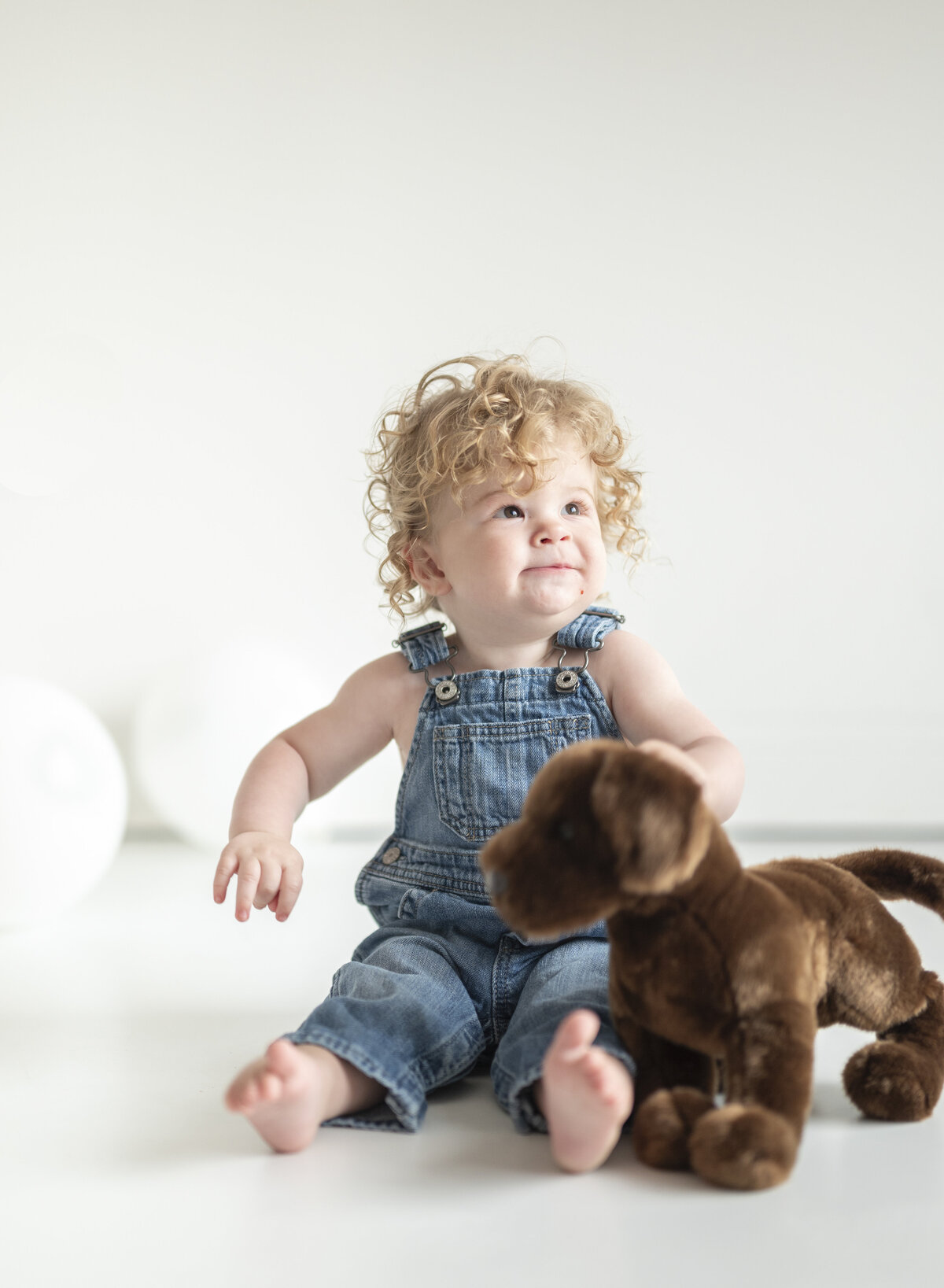 Toddler boy with curly hair in denim overalls sitting on the floor with a stuffed monkey, smiling brightly.