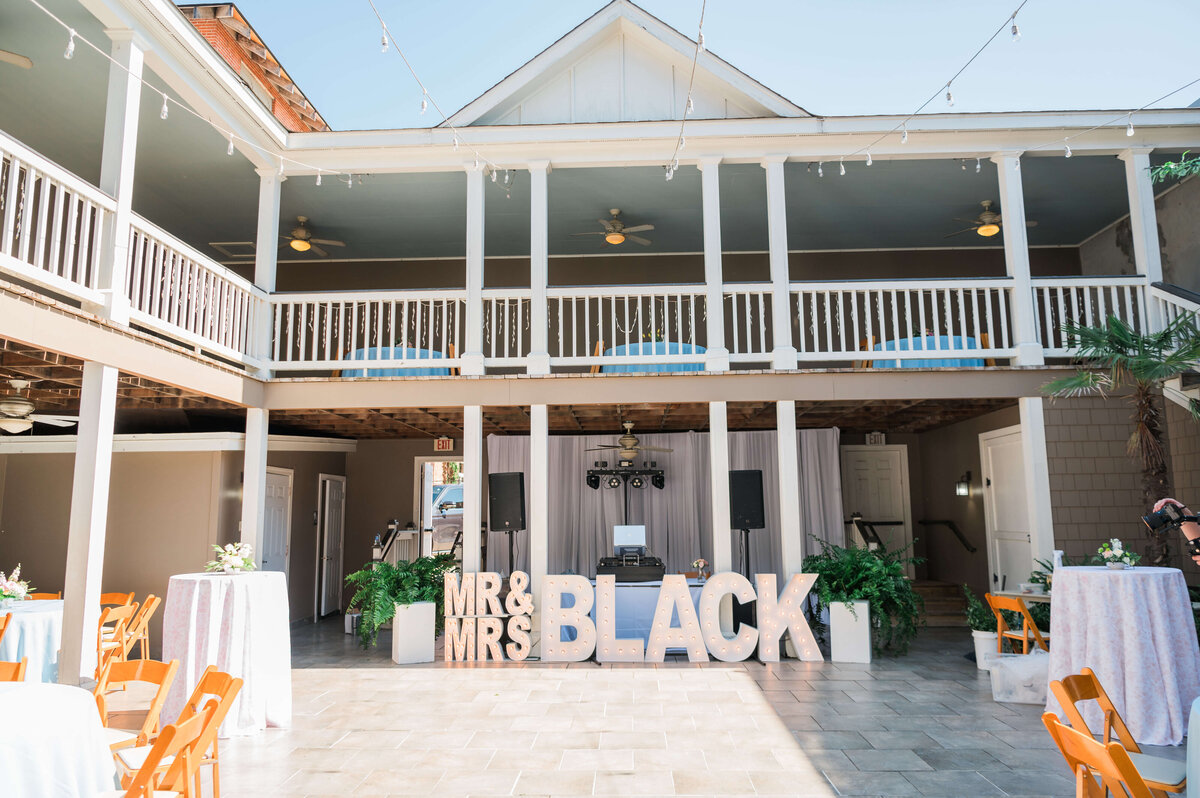 An outdoor courtyard set up for an event with round tables covered in light blue cloths, wooden chairs, string lights overhead, and large MR and MRS BLACK letters near a wall.