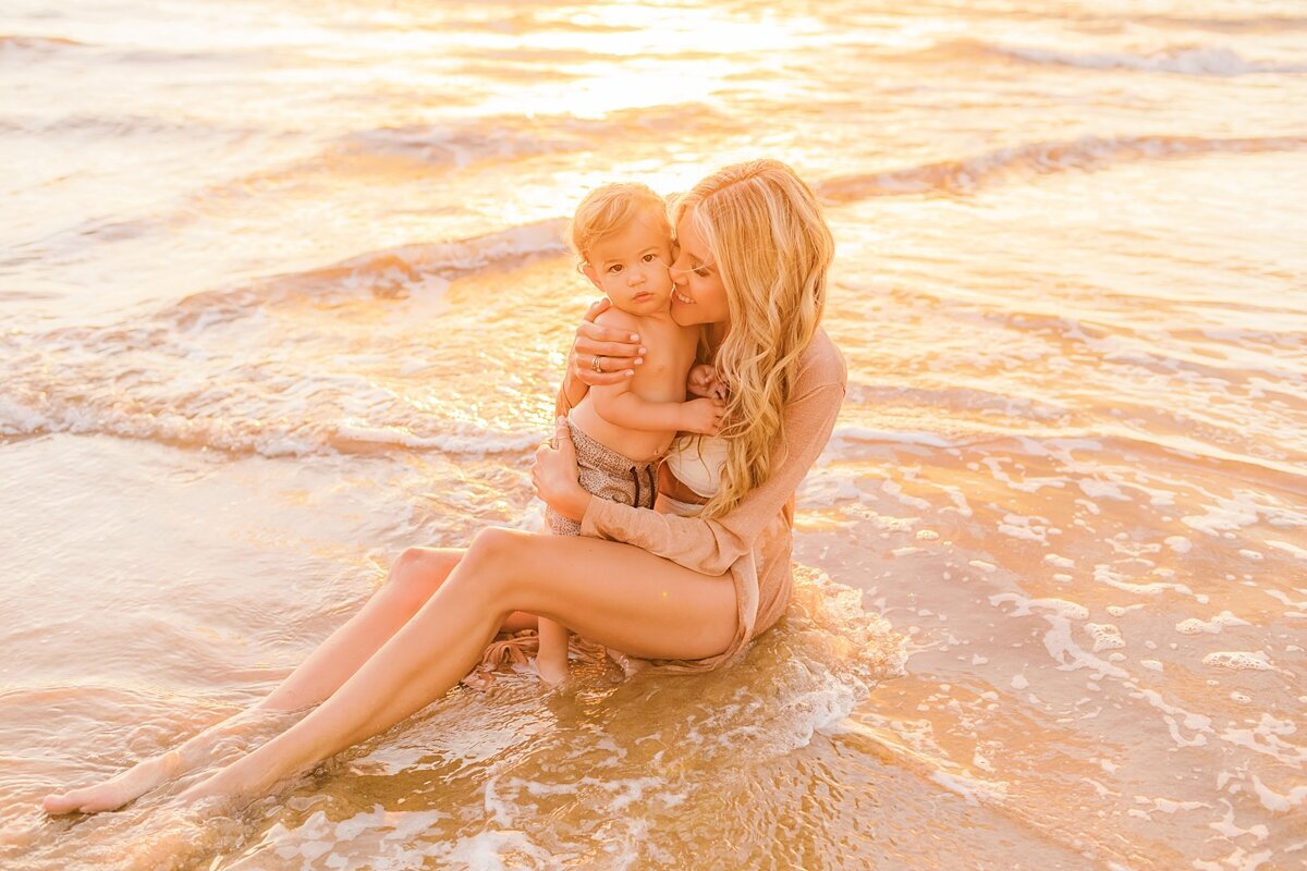 Mother with long blonde hair wearing a bikini cuddles her toddler on the shoreline next to the ocean in Maui