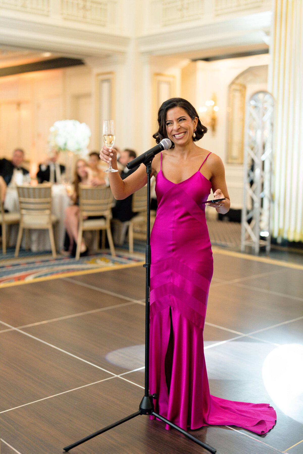 A woman in a vibrant magenta dress holds a microphone and a champagne glass while speaking on a stage. She is smiling, with seated guests and a bright, elegant interior in the background.