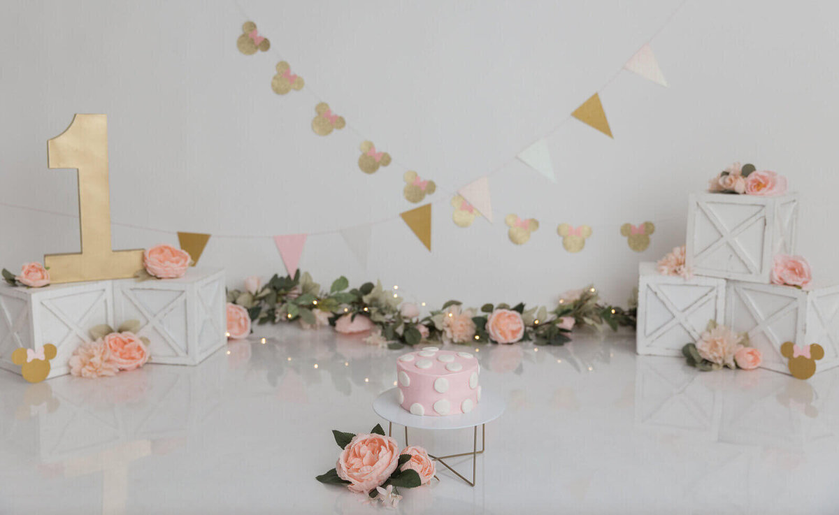 Disney-themed first birthday cake smash setup with a pink polka dot cake, gold Minnie Mouse decorations, and pastel-colored bunting.
