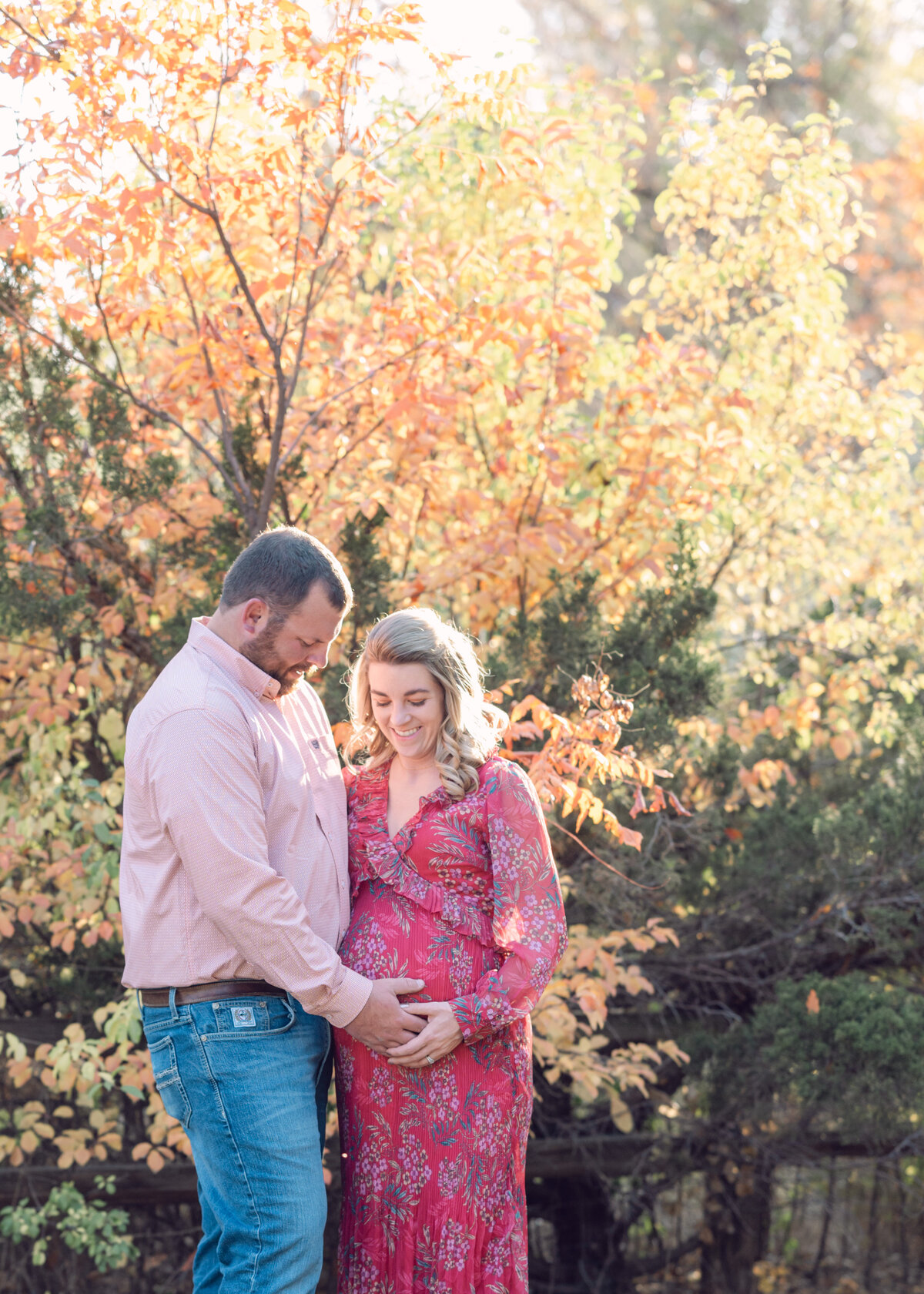 Cute couple smiles down at the wife's baby bump standing in front of a bright yellow tree in the fall landscape- photo by maegan r photography