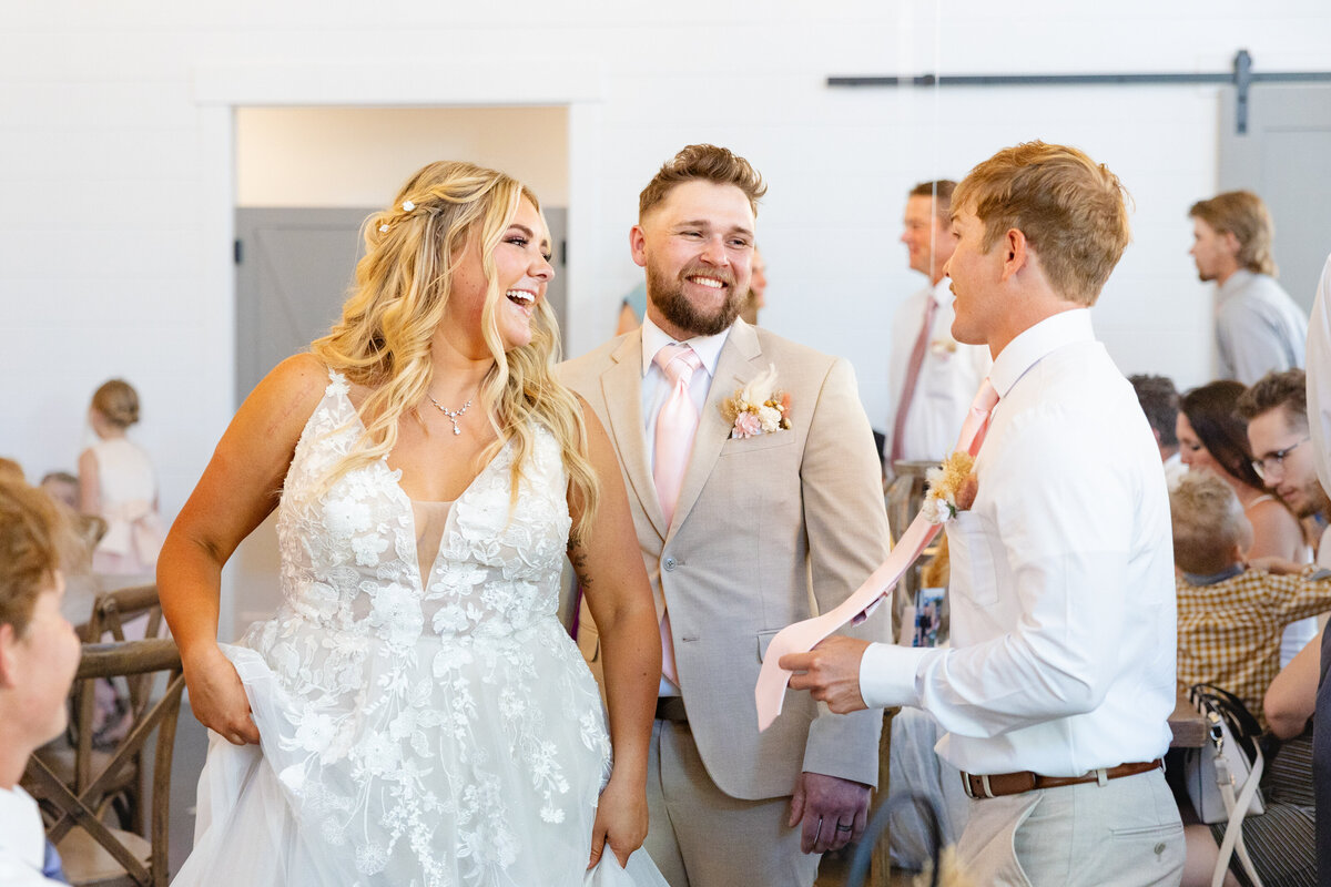 The bride and groom share a laugh with a guest at the wedding reception, with the bride playfully lifting her dress.