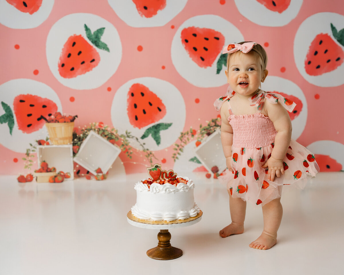 Adorable one-year-old baby girl celebrating her first birthday in a strawberry-themed photoshoot, wearing a cute pink dress adorned with strawberries. A strawberry cake sits in front of her as she beams with joy, captured by Yucaipa photographer Kaitlyn Dawn Photography.