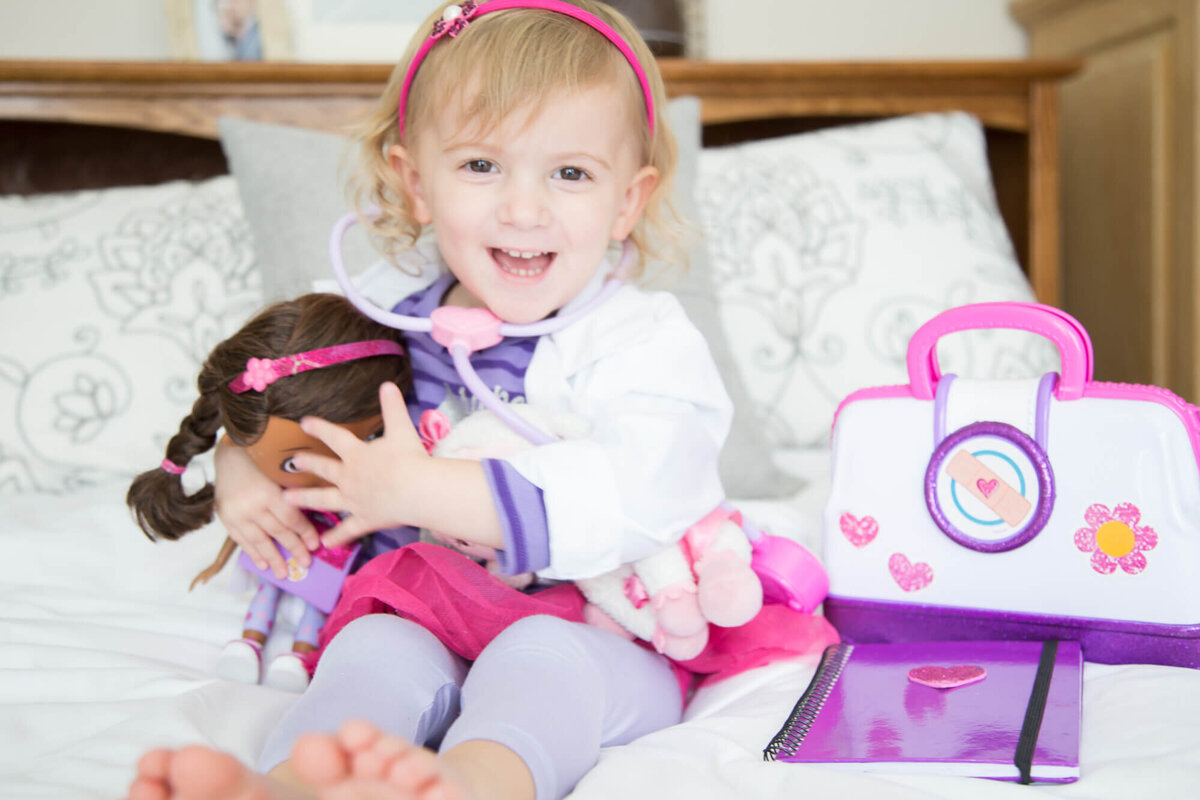 Happy blonde toddler girl smiling with her doll on a bed