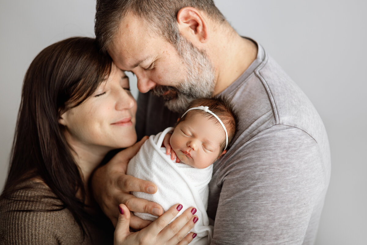 Newborn baby girl wrapped in white, held by smiling parents during a Grand Rapids, MI newborn photography session at Honeywild’s cozy studio.