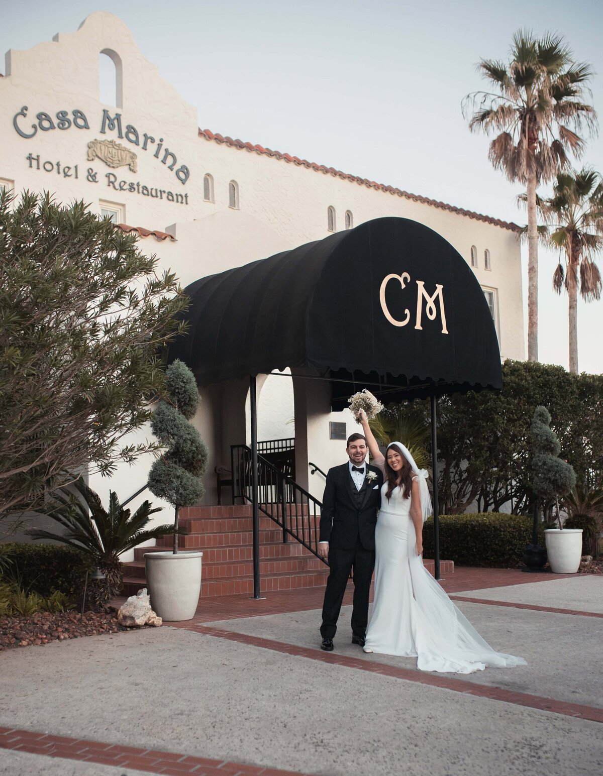  newlywed couple stands joyfully in front of the iconic Casa Marina Hotel & Restaurant in Jacksonville Beach, Florida. The groom wears a black tuxedo while the bride, in a sleek white gown, lifts her bouquet in celebration beneath the black CM canopy entrance. Palm trees and Mediterranean architecture create a timeless, coastal backdrop. Captured by a Jacksonville wedding photographer who specializes in elegant and true-to-color wedding moments at historic venues like Casa Marina.