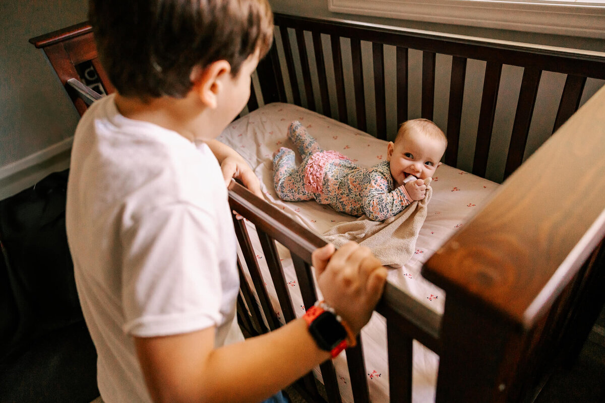 Infant girl is on her stomach in her dark wood crib, looking up at her big brother with a huge gummy smile.