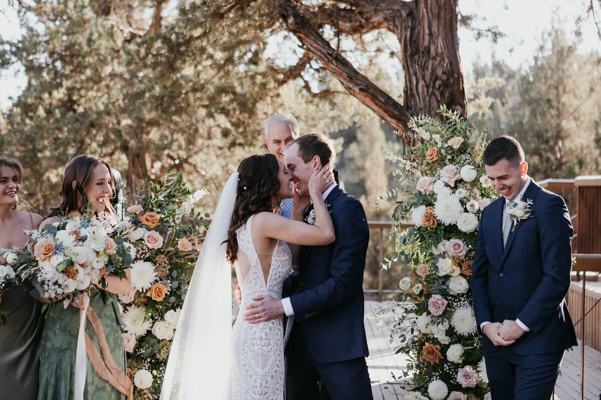 Bride and groom kiss each others during their wedding ceremony with floral pillars framing them.