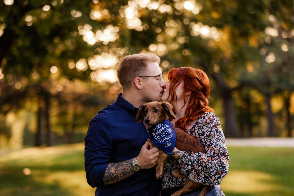 An engaged couple kisses while holding their pup.