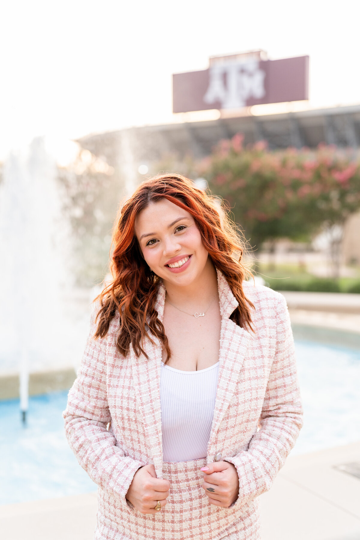 Texas A&M senior girl wearing checkered blazer and shorts and holding jacket while smiling in front of fountain and Kyle Field