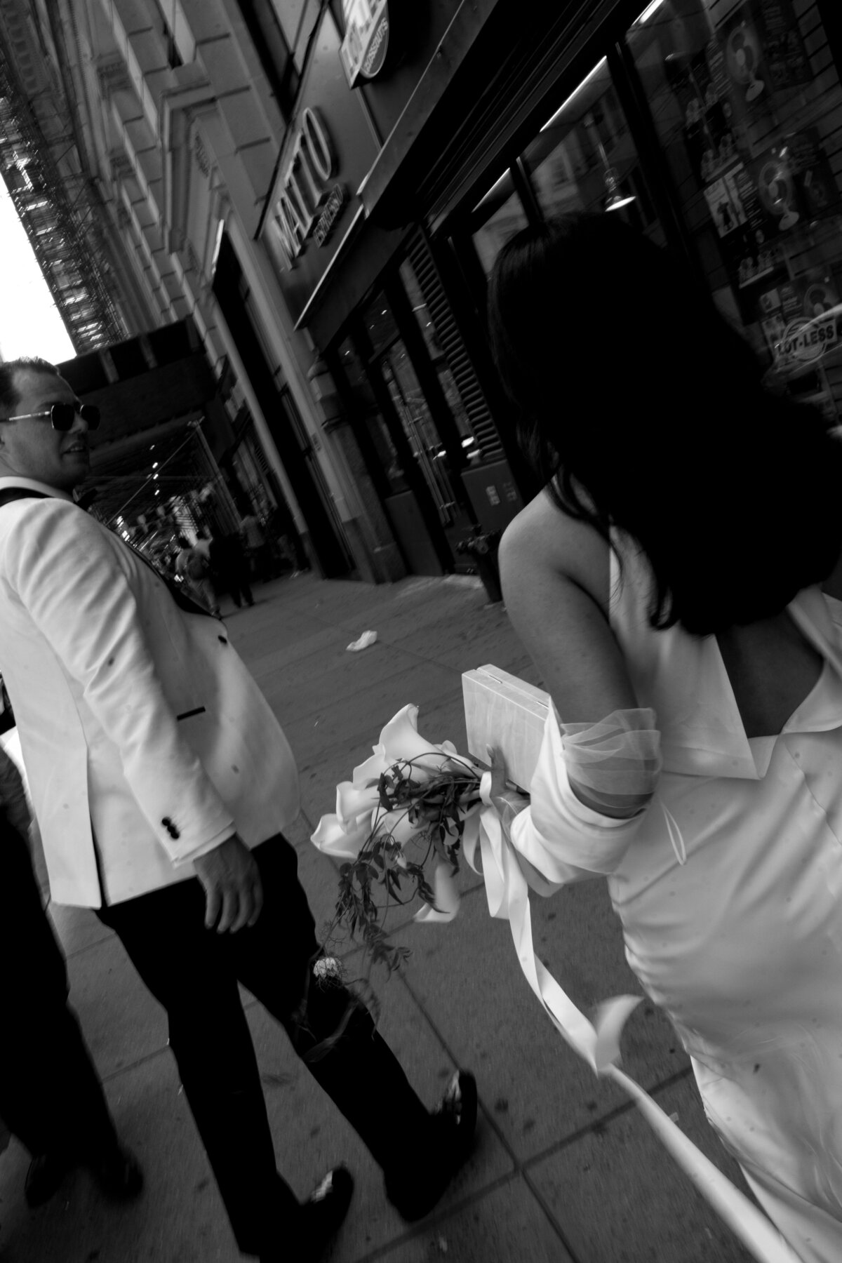 Black and white photo of a bride holding her bouquet while walking through the streets of New York City, her groom looking back at her during their City Hall elopement.