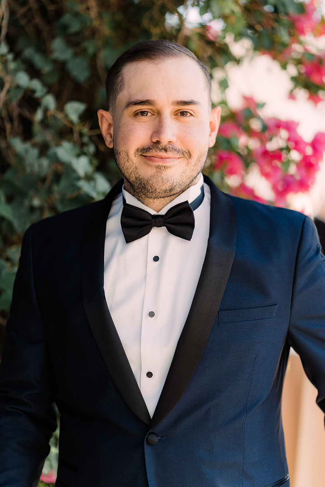 A portrait of the groom wearing a black tuxedo with a black bow tie, smiling with a backdrop of green foliage and bright pink flowers.