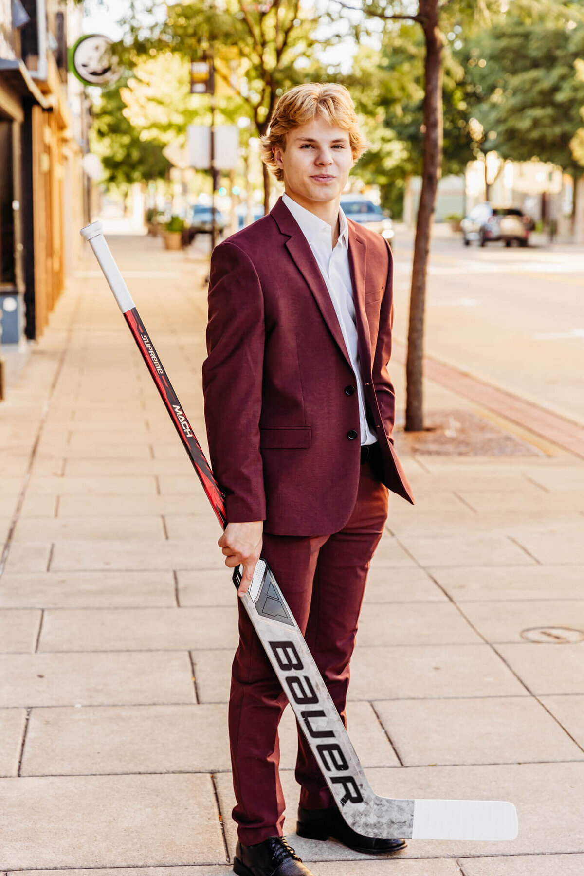 young man in a burgundy suit standing on a sidewalk in Neenah and holding his hockey stick with one hand.