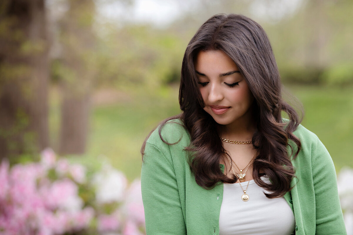 Senior close-up with dark wavy hair and green cardigan, surrounded by blooming azaleas in Charlotte NC.