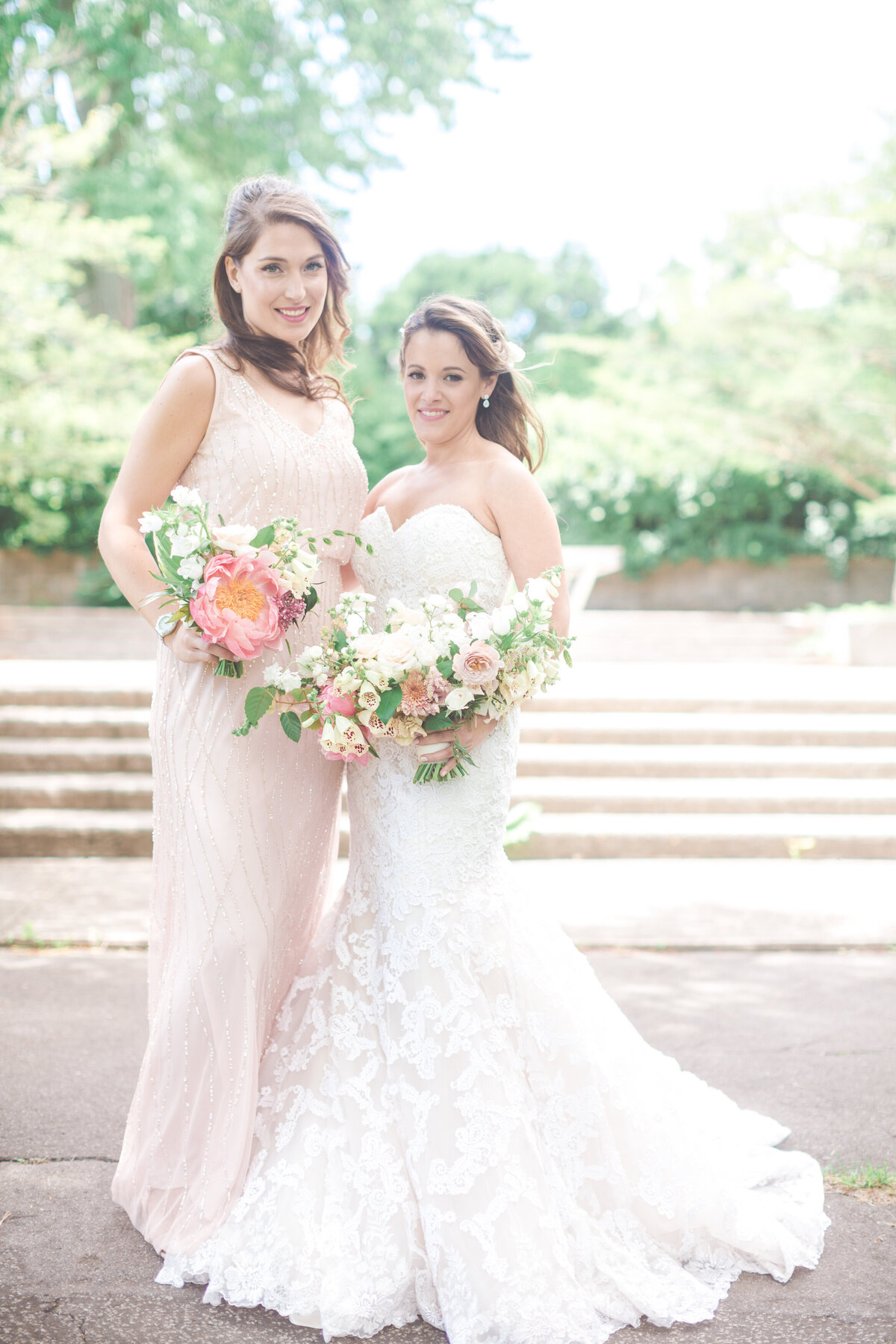A bride and bridesmaid smile in a Montreal park holding large bouquets of flowers.