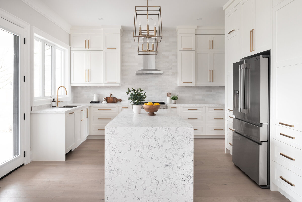 Kitchen design with all white cabinets, gold hardware, white and grey subway tiles, and a large marble waterfall countertop on the kitchen island at the centre.