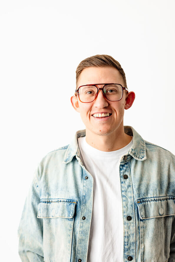 Man in jean jacket and glasses smiles at camera in a white photo studio.