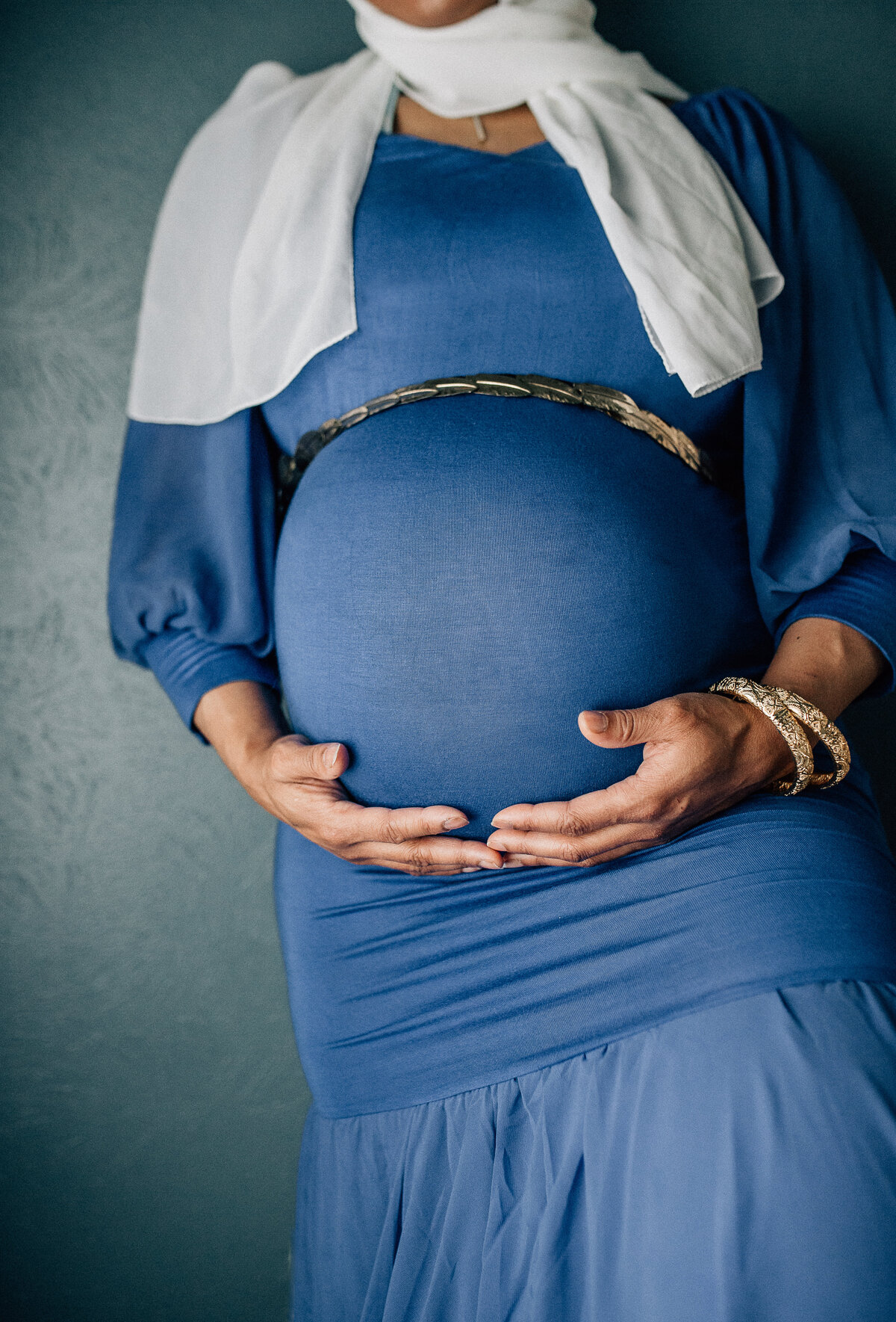 Maternity photoshoot with woman wearing blue dress, white scarf, and holding her belly.