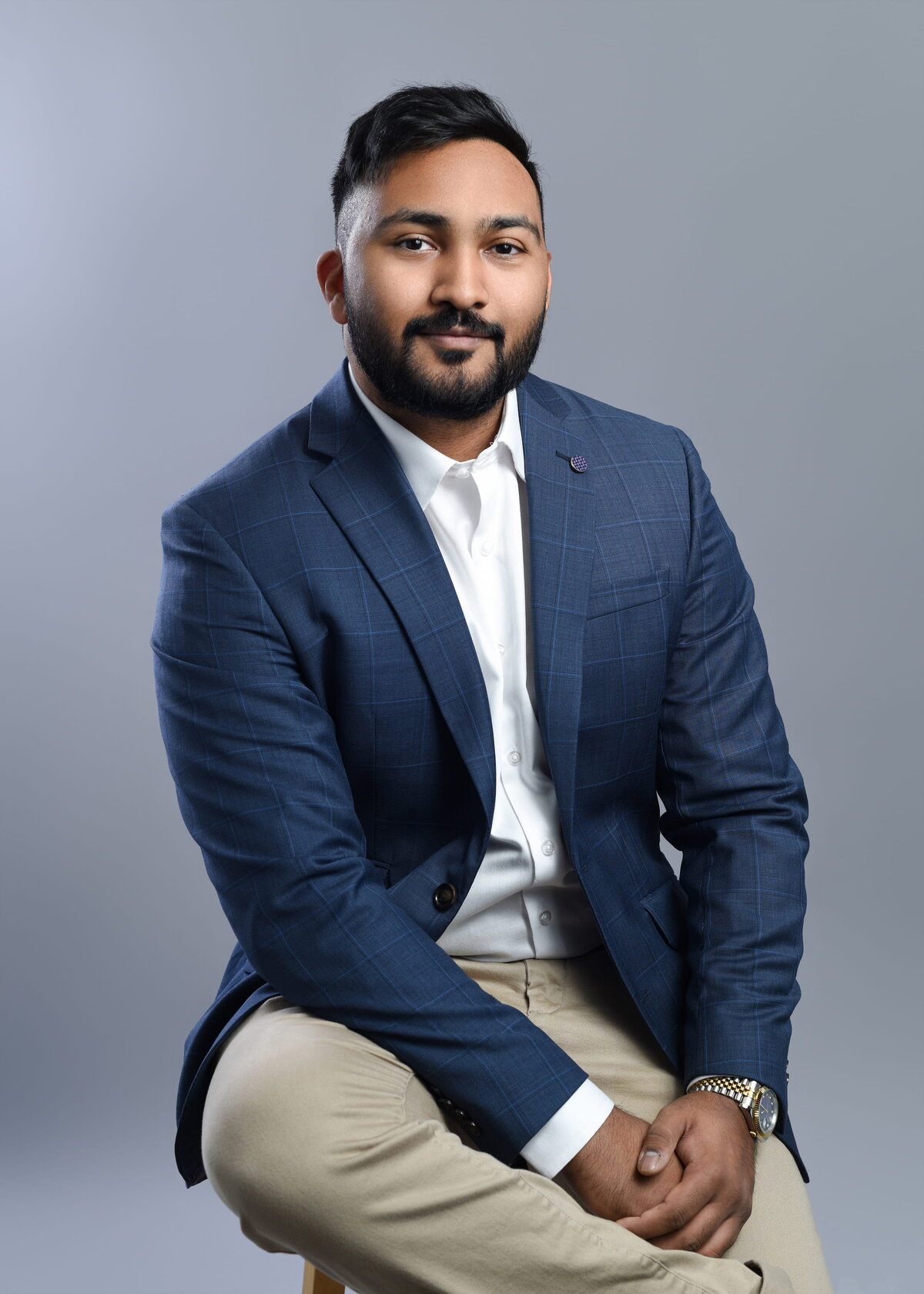 Professional headshot of an individual seated on a stool, wearing a blue checkered blazer, white dress shirt, and beige pants. The subject’s poised posture and confident expression against a plain background create a polished and approachable look—ideal for business branding and corporate photography